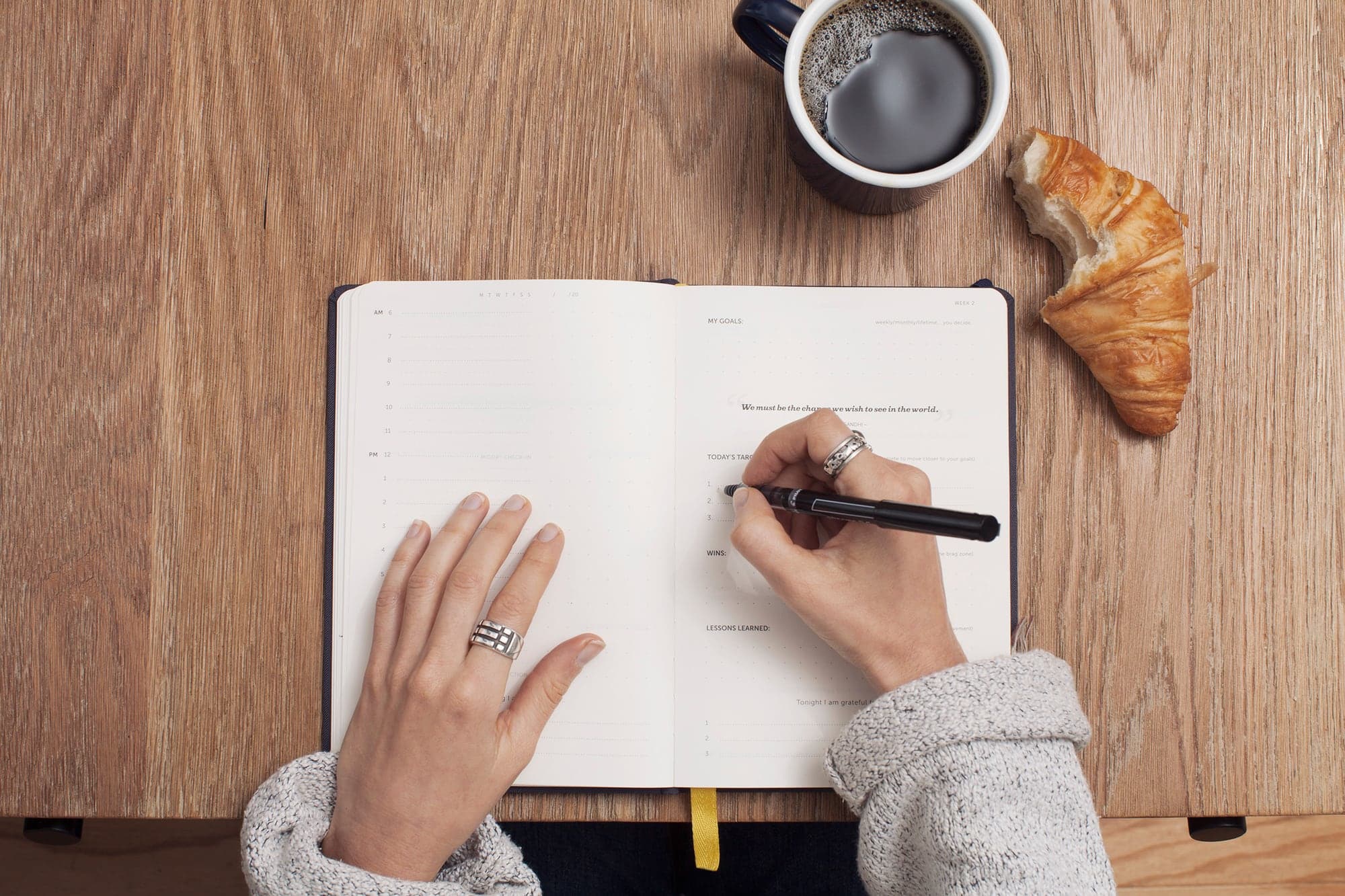 Deux mains écrivent dans un carnet ouvert, à côté d'une tasse de café et d'un croissant.