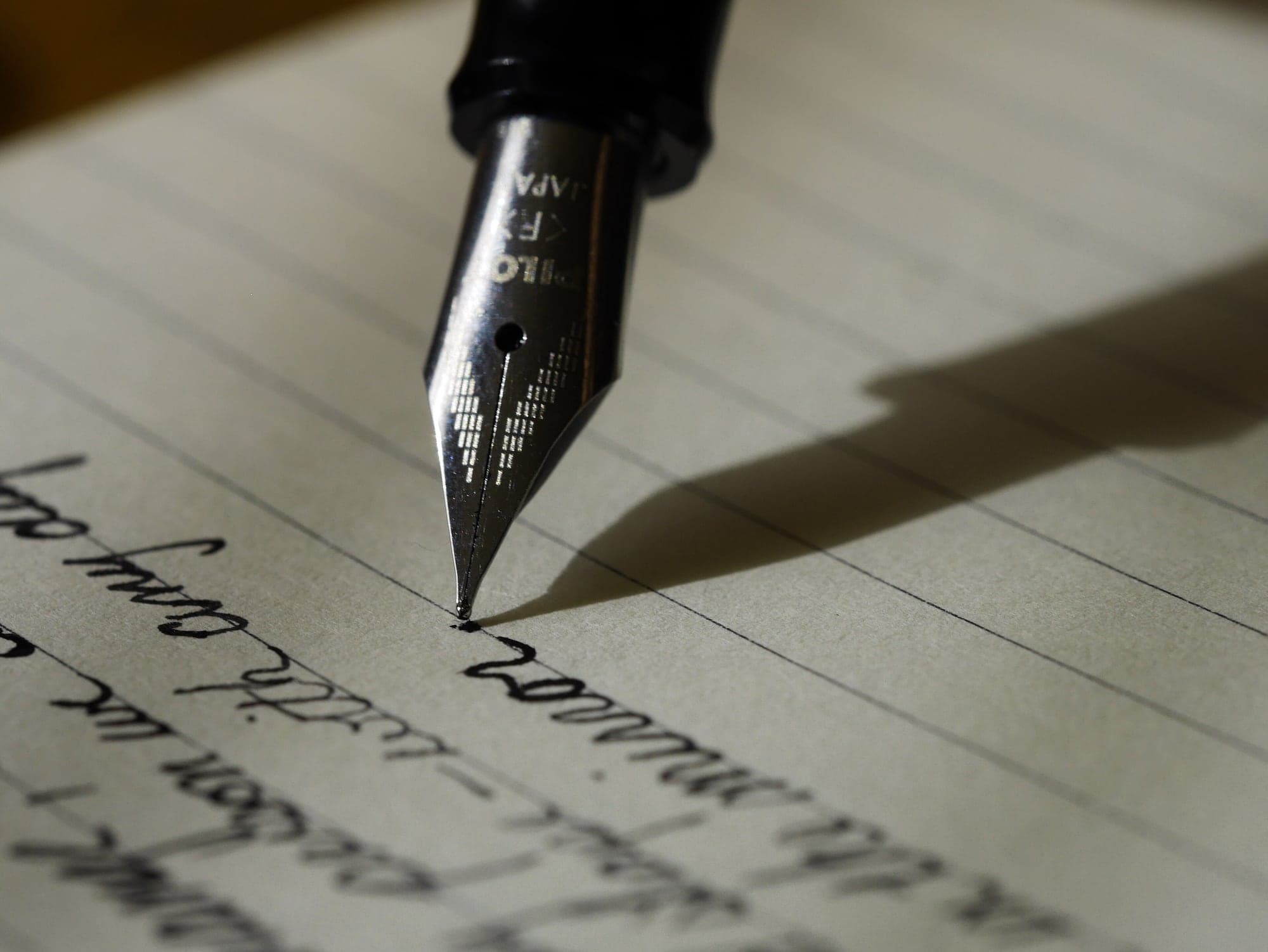 Une plume de stylo-plume trace une lettre manuscrite sur papier ligné, dans une lumière rasante.